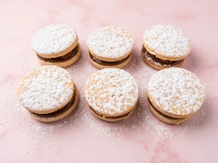Six alfajores dusted with powdered sugar on pink marble.
